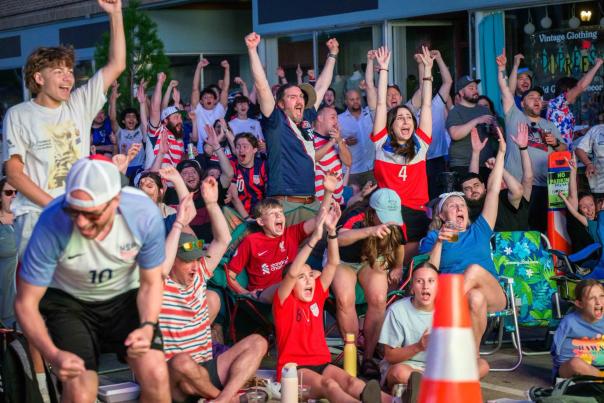 A crowd of soccer fans cheers with raised arms during an outdoor watch party on a city street.