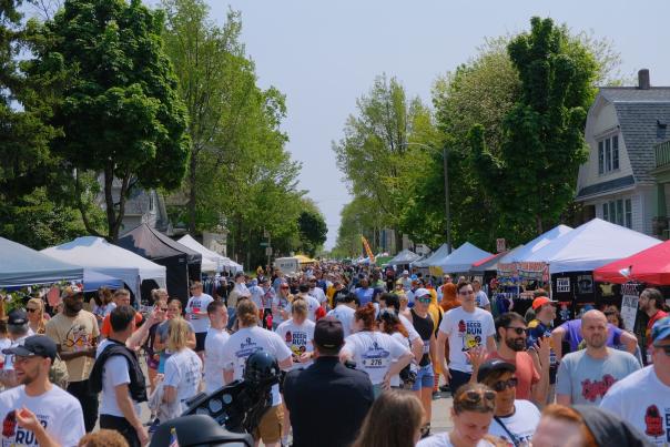 A crowded street festival stretches down a tree-lined residential block, with vendor tents on both sides and large groups of people walking and browsing. Many attendees wear matching “Locust Street Beer Run” shirts and race bibs, mingling among booths selling clothing, food, and drinks.
