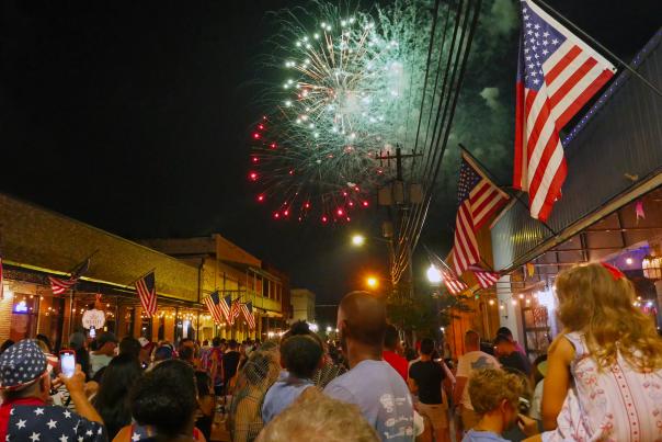 Fireworks light up the night sky over Olde Towne Slidell with a crowd below and American flags on display.
