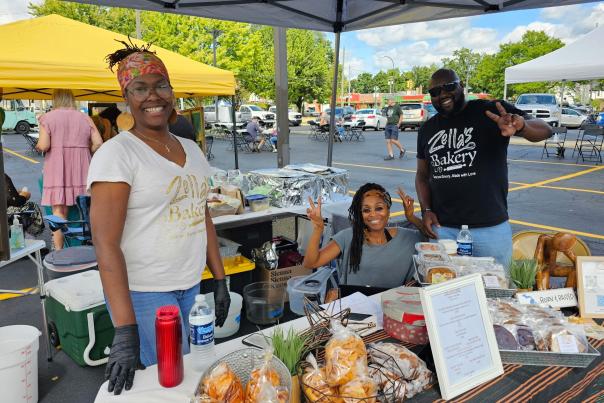 Three people pose behind a taple filled with baked goods for sale, set up in a parking lot. A person's T-shirt reads Zella's Bakery
