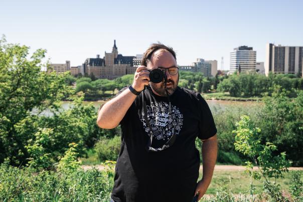 A man with a camera on a river bank with city in the background