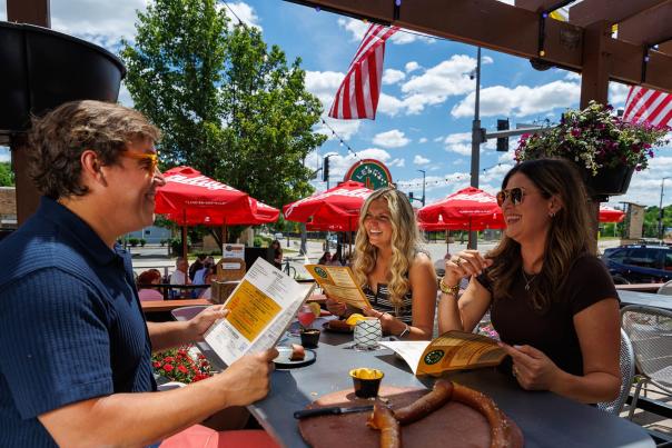 Two women and a man sitting at an outdoor patio table at Leff's Lucky Town, with bright red patio umbrellas and a blue sky visible in the background.