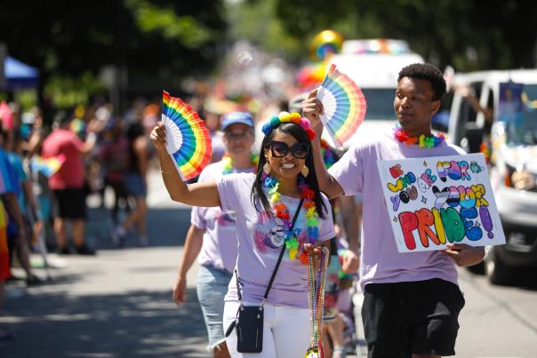 Two people in the 2025 Rochester Pride Parade with rainbow fans and a sign that reads "Be who you are for your PRIDE" in the colors of various Pride flags.