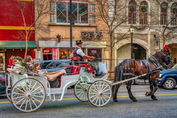 A white horse-drawn carriage decorated with Christmas garland and lights trots down Main Street in downtown Greenville, SC.