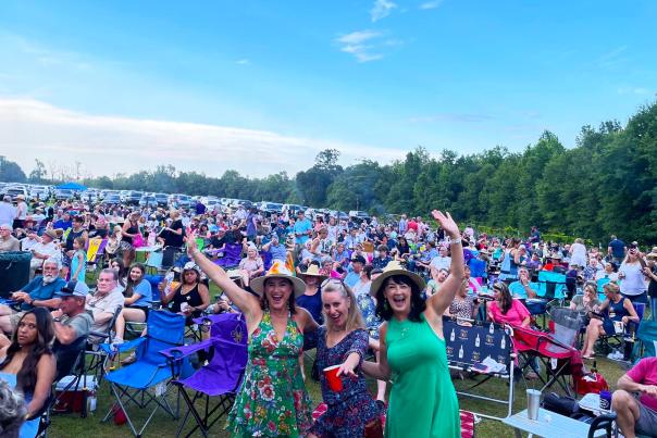 Large crowd seated in lawn chairs at Wild Bush Farm & Vineyard during a Jazz’n the Vines concert, with three women in the foreground smiling and raising their arms as guests gather on the grassy field under a clear evening sky.