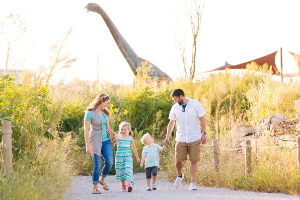 Family holding hands walking in front of a brontosaurus dinosaur in the background at Field Station Dinosaur