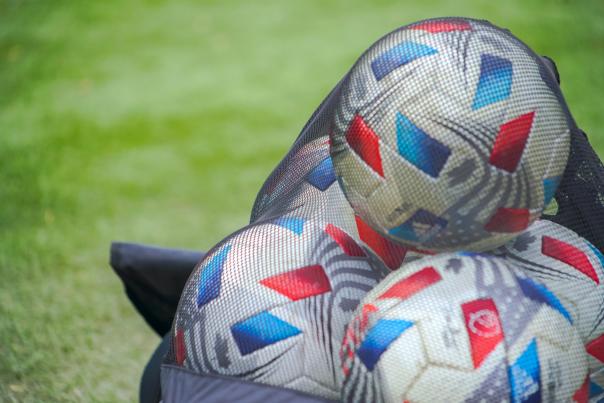 A net full of soccer balls rests on a bright green field. The soccer balls are mostly white with swaths of metallic red and blue.