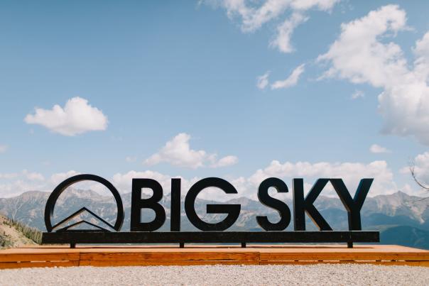 Black sign reading "BIG SKY" against a backdrop of mountains and blue sky with scattered clouds, promoting outdoor adventure and exploration.
