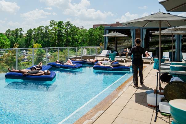 Six people relax on inflatable mattresses in the rooftop infinity pool at The Westin at The Woodlands. Beside the pool, a woman in black offers therapeutic words while a man plays singing quartz bowls, wind chimes, and a gong.