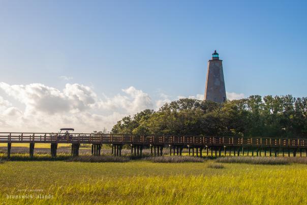 a golf cart crossing a wooden bridge over marsh with a stone lighthouse in the background