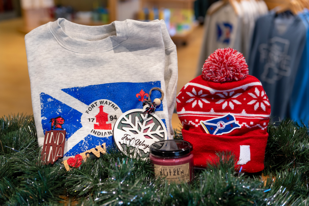 Collection of Fort Wayne merchandise, including a winter hat, sweatshirt, christmas ornament, and candle, on a table with holiday gardland