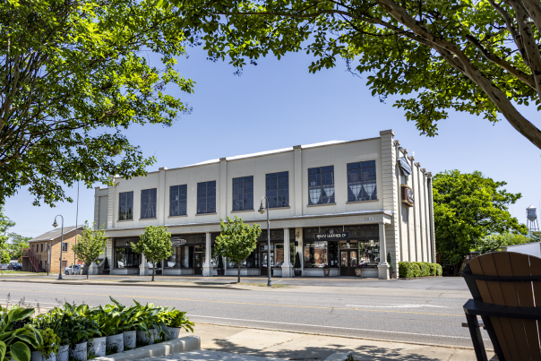 white building facing a street with three retail store fronts on a bright sunny day with green trees and plants framing the foreground.