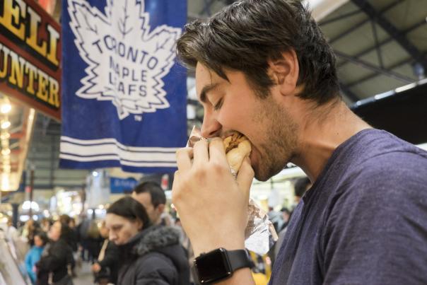 A man eating a peameal bacon sandwich with a Toronto Maple Leafs flag behind him
