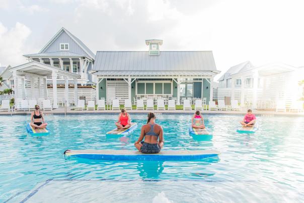 A woman with crossed legs sits atop a surf board floating in a pool, back to the camera. Four women, also with crossed legs, sit atop surf boards facing the instructor.