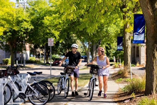 An Asian man and Asian woman walk electric bikes to their docking station on a street surrounded by lush greenery