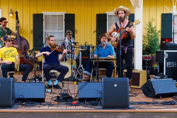 A band performs on an outdoor stage at the Abita Springs Busker Festival, with musicians playing guitar, fiddle, upright bass and drums in front of a yellow building.
