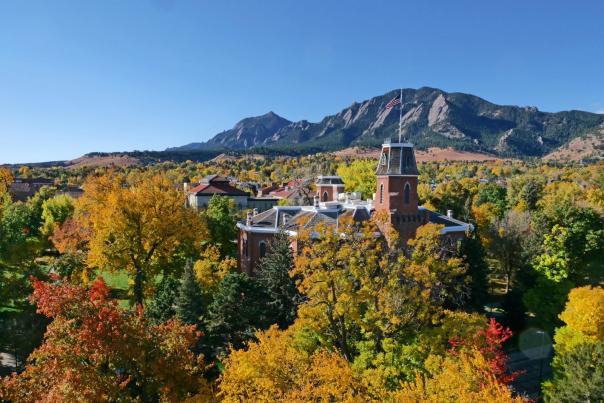 Aerial view of the University of Colorado Boulder campus framed by vibrant fall foliage with the Flatirons rising in the background under a clear blue sky.