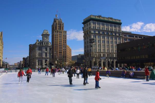 People skate on the outdoor ice rink in Clinton Square on a sunny winter day, with historic downtown Syracuse buildings rising in the background and groups of visitors gathered along the edges of the rink.