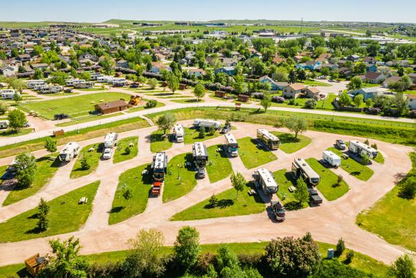 Aerial view of an RV park on a bright, sunny day, with neatly arranged RVs on grassy lots and a suburban neighborhood in the background.