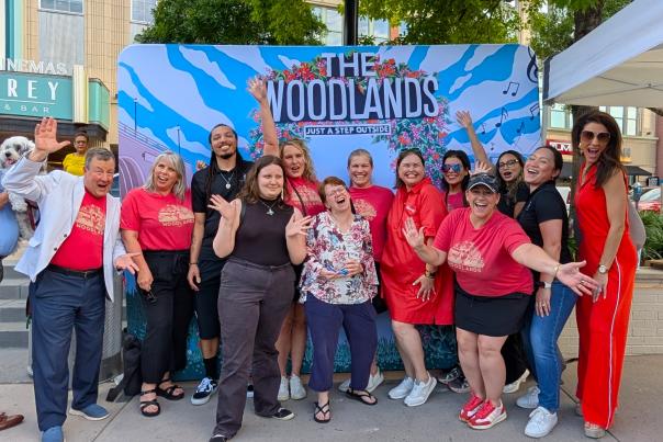Thirteen people smile and pose for the camera in front of a backdrop of a mural of The Woodlands. Among them are Visit The Woodlands Staff Members: Nick Wolda, Julie Quinn, Josie Lewis, Ashley Fenner, Elizabeth Eddins, Carla Twidell, and Amber Parsley. Others include Market Street staff, winners of the National Travel and Tourism Week Awards, a local FOX News anchor, and more.