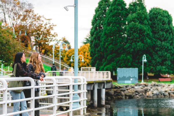 Two people pause along Nanaimo’s waterfront walkway, leaning on a railing and enjoying the view of the calm water and nearby park.