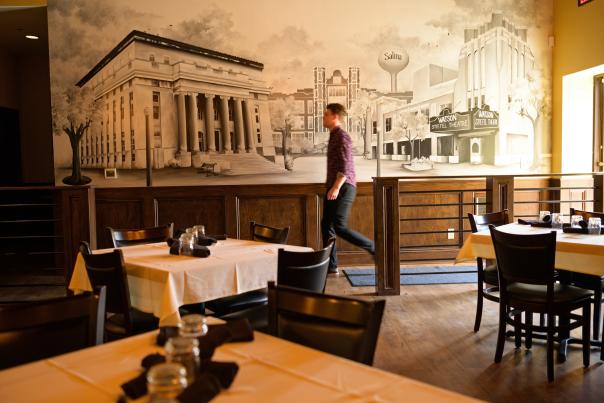 Inside of a restaurant with linen covered tables. A man walks in front of mural on a wall.