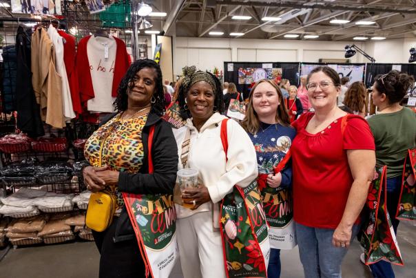 A group of smiling shoppers pose with festive Holly Day Fair tote bags while exploring vendor booths filled with holiday gifts and apparel at the Crown Expo Center in Fayetteville, NC.