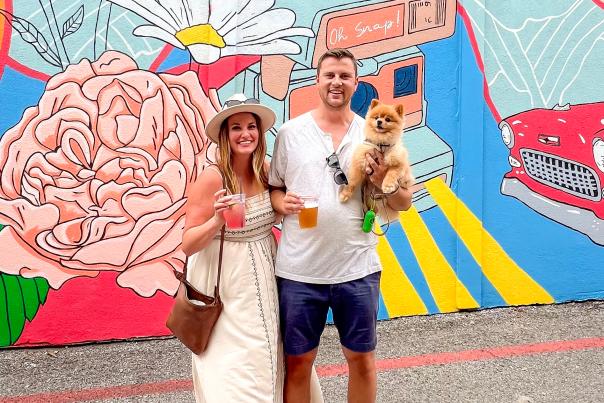 You couple - blonde woman in hat and man with beard, holding a small dog, standing in front of a colorful mural