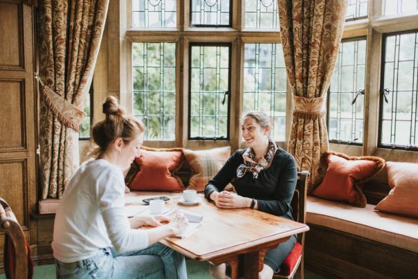 two women sit across from each other at a table in a bay window with cups of coffee smiling
