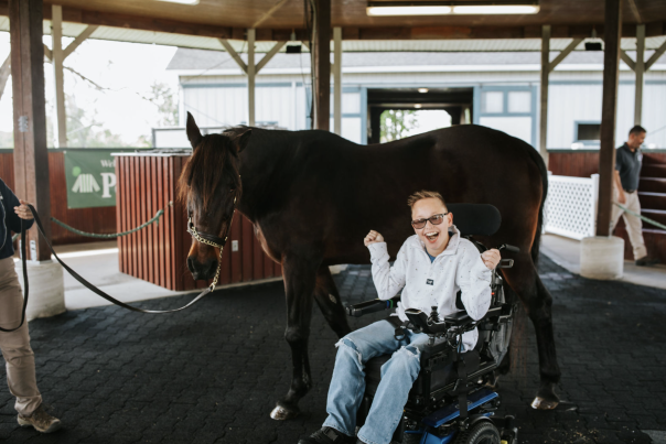 Boy in motorized wheelchair smiles in front of horse.