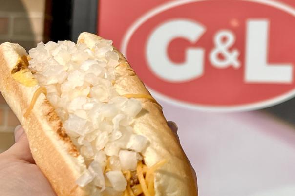 A hot dog topped with cheese and onions, held in front of a G&L sign.