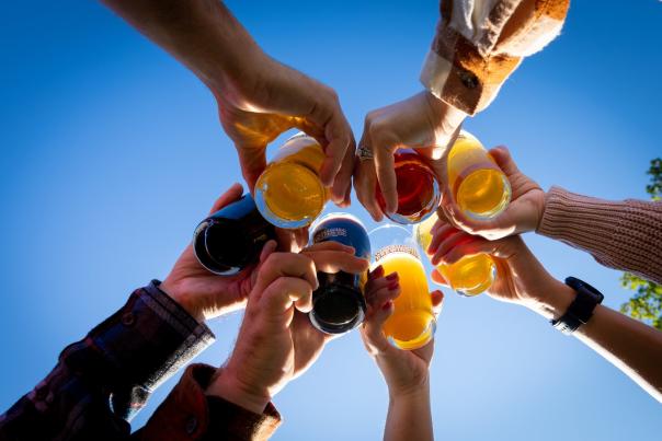 A view of seven people cheersing beer from the ground up with a bright blue sky in the background.