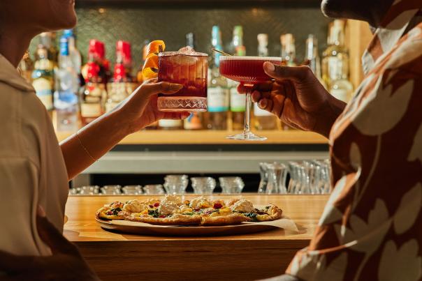 A couple shares drinks over a pizza at the True Food Kitchen bar. The handcrafted pizza features a thin crust, spinach, peppers, and large balls of melted cheese.