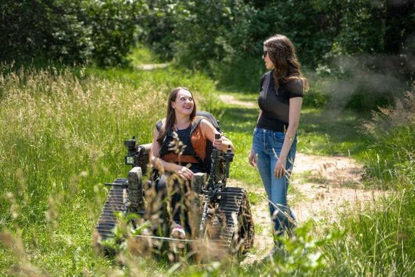 Two women are on a grassy woodland path, one seated in a wheelchair with tracks, smiling, while the other stands beside her. The scene is bright and conveys happiness.