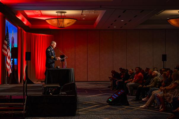 Palmer Buck, Chief of The Woodlands Fire Department, speaks at the Texas State Association of Fire Fighters 49th Biennial Convention. Buck is in uniform, standing at a clear podium on a makeshift stage backlit in red, and his image appears on a large projection screen to the right of the frame. A crowd listens intently.