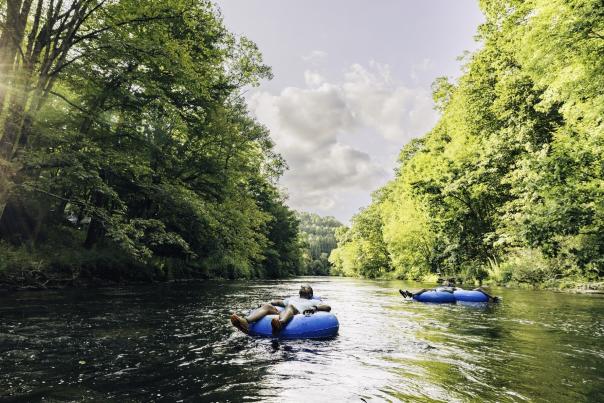 A calm river with lush green trees on either side has three people in floats, the closest of which has their eyes closed looking up at the sky.