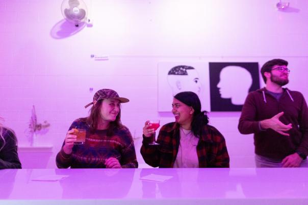 Two women look at each other while sitting at a bar and drinking cocktails at a bar with purple lighting and very contemporary art
