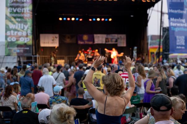 A white woman raisers her hands in as she dances in a crowd in front of a stage where music is being performed at La Fete de Marquette