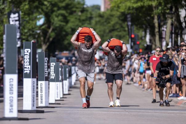 Two men hold orange sandbags on their back and walk up State Street in Madison for the CrossFit Games.