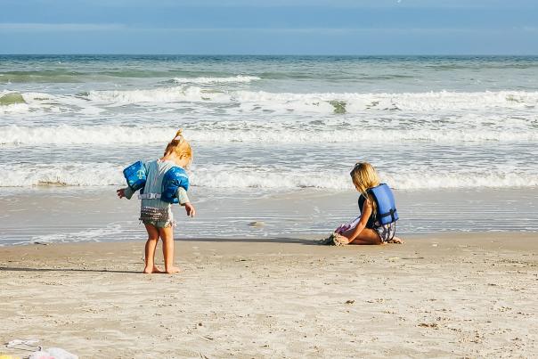 Two blonde toddlers, one in water wings and one in a life jacket, play on the beach by the water. one is kneeling in the sand, one is standing, walking towards the water