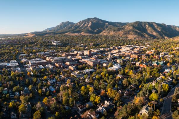 Aerial view of downtown Boulder surrounded by colorful fall foliage, with the Flatirons and forested foothills rising in the background beneath a clear blue sky.