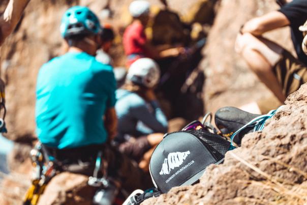 A black ballcap shows the Colorado Mountain Club words and logo in the foreground with blurred rock climbers behind gathering at the climbing area of North Table Mountain in Golden CO.