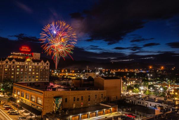 Fireworks burst in the night sky above downtown Rapid City, with the illuminated Hotel Alex Johnson and city lights glowing beneath dark clouds at dusk.