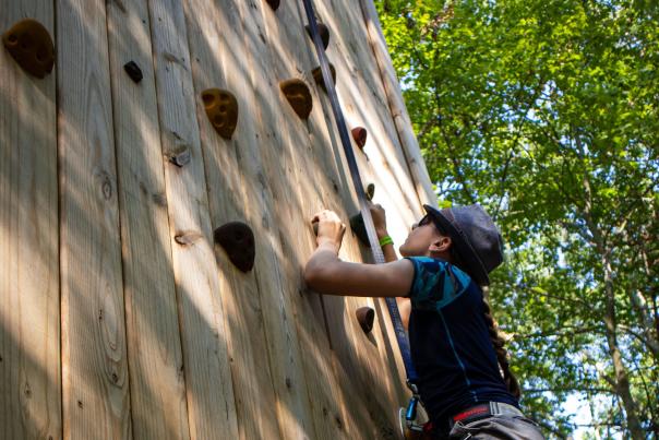 A young climber ascends a wooden outdoor climbing wall surrounded by forest at Muskegon’s Luge Adventure Sports Park.