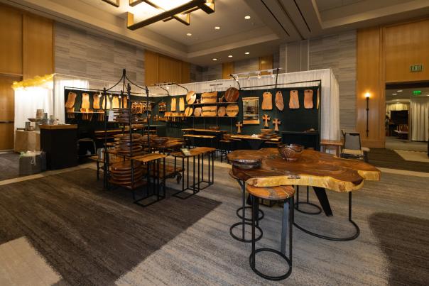 A craft show booth displays elaborate wood crafts. Sleek, organically-shaped cutting boards hang along the back wall of the booth. A tiered display in the middle of the booth shows off large mixing bowls and Lazy Susans. A large wooden breakfast table with matching barstools are also shown for sale.