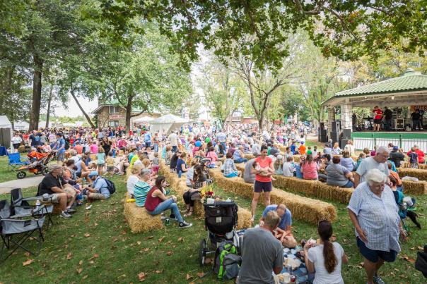 A crowd of people gathered on a grass lawn and sitting on hay bales, waiting for a show to start on a stage