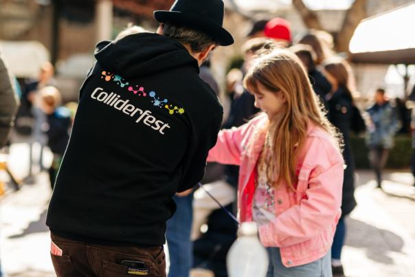 Man in a black Colliderfest hoody shows a science trick to a young girl