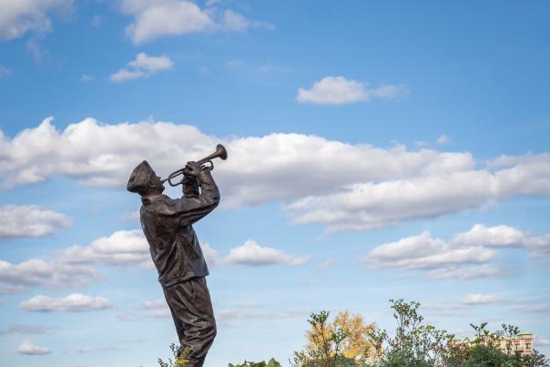 Statue of a person playing a trumpet, set amidst a vibrant garden of multicolored flowers and plants, under a partly cloudy sky.