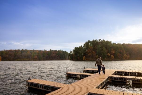 An adult and a small child walk along a wooden dock extending over a lake, with rippling water, autumn-colored trees, and a wide blue sky in the background.