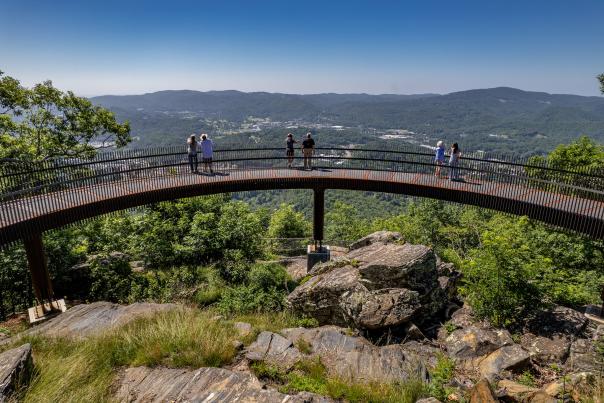 A curved, metal walkway stands on a rocky mountain peak surrounded by green trees, with three couples taking in the views of a mountainous horizon and the Town of Boone below.